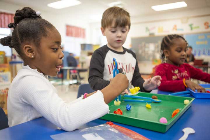 Three children play a game with tongs in a preschool classroom