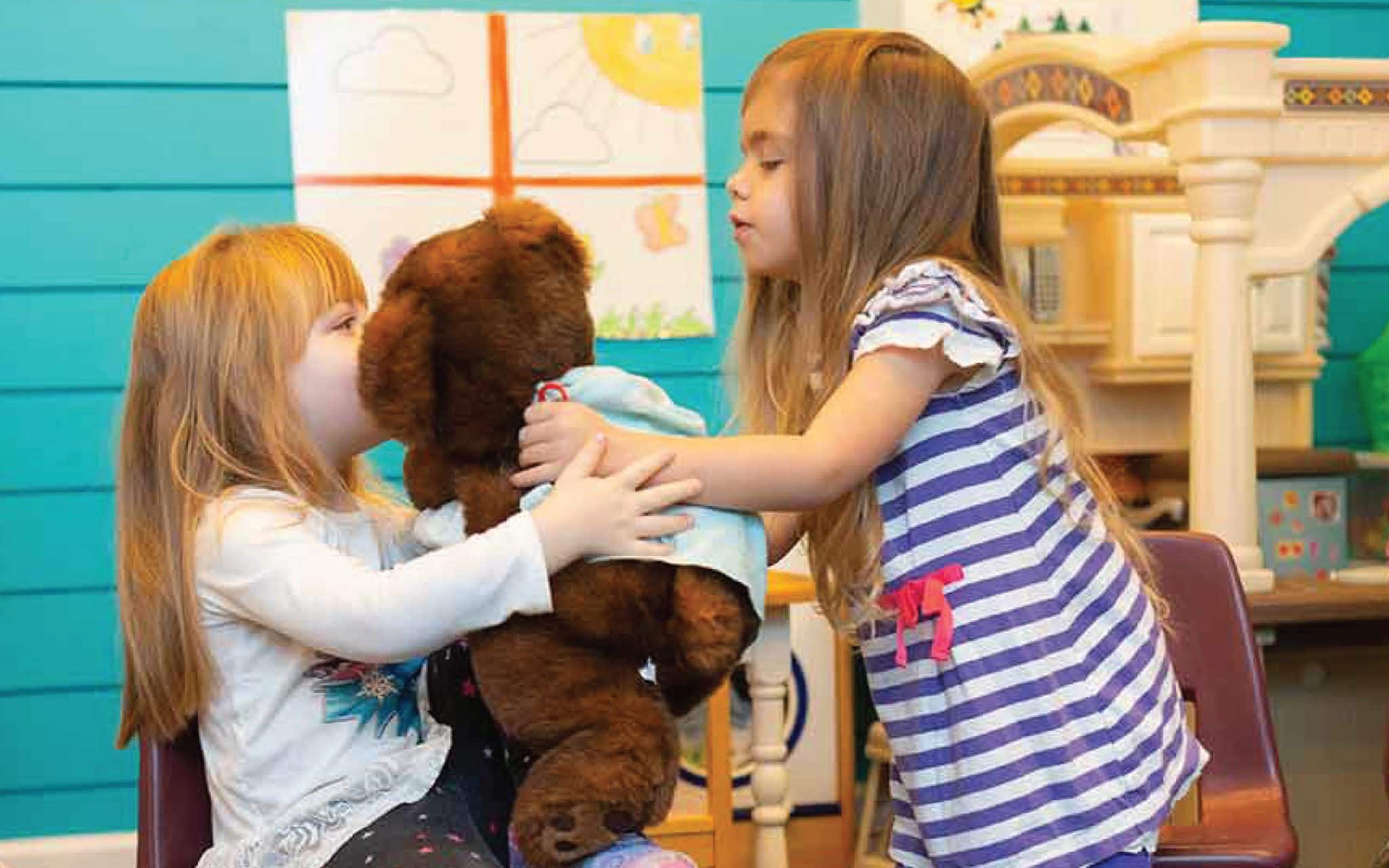 One girl hands a teddy bear to another girl who is seated in a preschool classroom