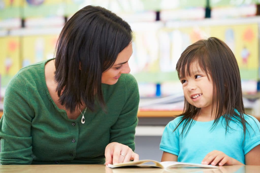 Elementary Pupil Reading With Teacher In Classroom Looking At Each Other Smiling